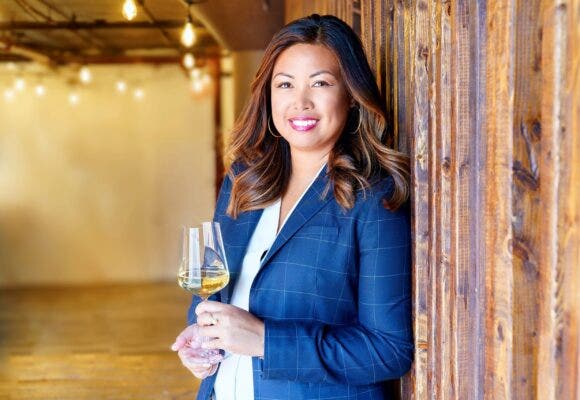 Wine Enthusiast tasting director Anna-Christina Cabrales holds a glass of white wine standing against a wooden wall
